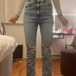 Högmidjade ljusblå jeans från Zara - Säljer ett par ljusblå jeans från Zara i storlek 34. Modellen är högmidjad med raka ben/bootcut och har snygga slitningar vid knäna. Perfekta för dig som gillar en avslappnad och cool stil. Jag är 174 och dom går hela vägen ner till golvet, perfekt för alla långa tjejer 🩷