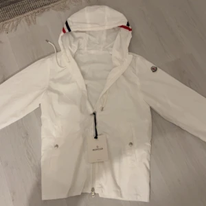 Vit vindjacka från Moncler med huva - Snygg vit vindjacka från Moncler med huva och dragkedja framtill. Jackan har Moncler-logga på ärmen och coola ränder i rött och blått på huvan. Två fickor med knappar och justerbar snörning vid huvan. Perfekt för dig som gillar stilren och sportig look. Märket går att scanas👍M som också passar S 
