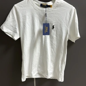 Vit t-shirt från Polo Ralph Lauren - Vit t-shirt från Polo Ralph Lauren med klassisk rund hals och korta ärmar. T-shirten har den ikoniska broderade loggan i svart på bröstet och är tillverkad i mjuk bomull. Perfekt basic med stilren design och snygg passform.