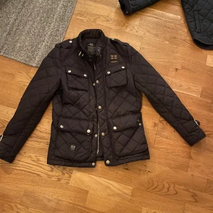 Mörkblå quiltad jacka från Morris  - Quiltad mörkblå jacka från Morris med fyra fickor framtill, silverfärgade knappar och dragkedja. Snygga detaljer som patch med brittisk flagga och mönstrat innerfoder. Jackan har lång ärm och justerbara spännen vid handlederna. Perfekt för dig som gillar stilren look. Storlek XS men passar mer som S. Skick 10/10