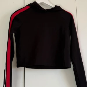 Svart croppad hoodie med röd och vit rand - Svart croppad hoodie med långa ärmar och huva. Snygga röda och vita ränder längs ärmarna ger en sportig vibe. Materialet är mjukt och känns som bomull eller polyester. Perfekt för dig som gillar streetstyle och vill sticka ut med coola detaljer.
