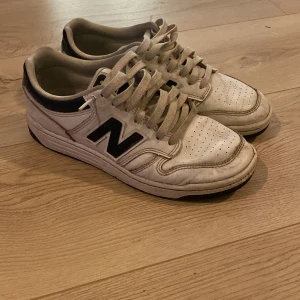 Vita New Balance sneakers med svart detalj - Säljer ett par klassiska New Balance sneakers i vitt skinn med svarta detaljer och stor N-logga på sidan. Skorna har snörning, perforerad tå och platt sula. Perfekta för dig som gillar retrostil och vill ha bekväma sneakers till vardags.