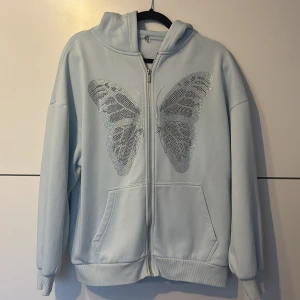 Ljusblå hoodie med fjäril och dragkedja - Snygg ljusblå hoodie med huva och hel dragkedja framtill. På bröstet sitter ett stort fjärilsmotiv i silverpaljetter som ger extra bling. Tröjan har långa ärmar, ribbade muddar och två praktiska fickor framtill. Perfekt för dig som gillar detaljer och chill stil.