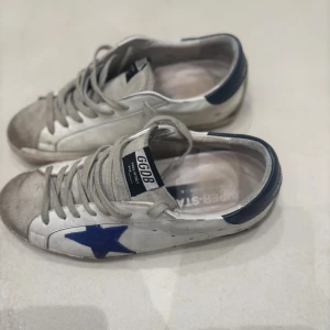 Golden Goose vita sneakers med blå stjärna - Golden Goose sneakers i vitt skinn med grå mockadetaljer och blå hälkappa. Sidan pryds av en blå stjärna och skorna har klassisk snörning. Snygg, avslappnad look med rund tå och platt sula. Perfekta för dig som gillar stockholmstil,grisch  och unika detaljer.