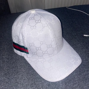 Vit keps från GG  - Säljer en vit keps  med klassiskt GG-monogram över hela kepsen. Kepsen har justerbar rem bak, gröna och röda ränder på sidan och är tillverkad i bomullsmix. Snygg och stilren accessoar för dig som gillar exklusiva märken. Väldig sällsynt 