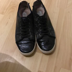 Svarta sneakers från Lanvin i skinn - Svarta sneakers från Lanvin med klassisk design, rund tå och svarta skosnören. Ovandel i glansigt skinn och vit platt sula. Metallöljetter och diskret logga på innersulan. Perfekta för en stilren och clean look.