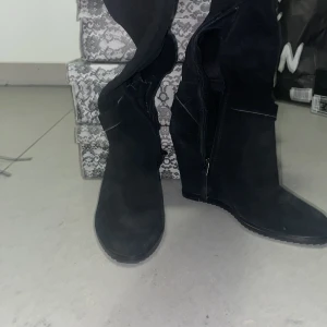 Svarta kilklacksstövlar i mocka - Snygga svarta stövlar med kilklack och högt skaft, tillverkade i mocka. Stövlarna har dragkedja på insidan och ett dekorativt spänne runt vristen. Perfekta för dig som vill ha en stilren look med lite extra höjd. Pris kan diskuteras