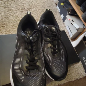 Svarta sneakers från KAYOBA strl 43 - Svarta sneakers från KAYOBA med vita detaljer och mönstrade inslag på ovandelen. Skorna har snörning oh en sportig, modern look med platt sula och rund tå. Perfekta för dig som gillar stilrena och bekväma skor till vardags eller träning.