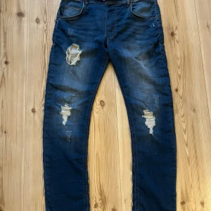 Blå ripped jeans Adrian Hammond strl 32 - Snygga blå jeans från Adrian Hammond i modellen Ohio Jogger. De har slitna detaljer och hål på framsidan för en cool, trendig look. Materialet är en mix av bomull, polyester och elastan som ger skön stretch. Klassisk femficksdesign och normal passform.