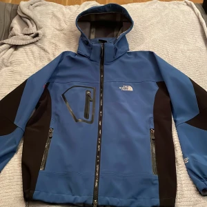 Blå vindjacka från The North Face L - Blå och svart vindjacka från The North Face Summit Series med huva och flera dragkedjeförsedda fickor. Jackan är tillverkad i Gore-Tex och har justerbara ärmslut. Perfekt för utomhusaktiviteter och har en sportig look.