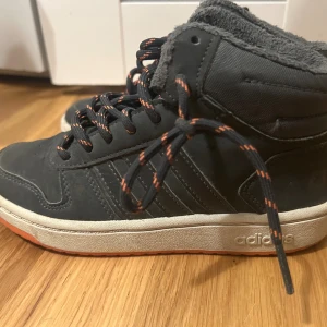 Adidas skor för barn  - Säljer ett par  adidas sneakers med högt skaft och orange detaljer på snörena. Skorna har mjuk, grå fleecefodring och klassiska adidas-ränder på sidan. Yttersulan är vit med orange undersida. 