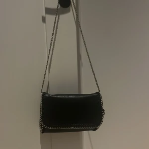 Svart axelväska med kedjedetaljer - Snygg svart axelväska i skinnimitation med silverfärgad kedja som axelrem och runt kanterna. Väskan har lock med tryckknapp och flera fack inuti, inklusive ett med dragkedja. Perfekt för dig som gillar en stilren men edgy look.
