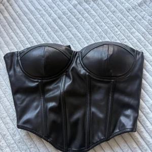 Svart skinnkorsett med kupor - Säljer en svart korsett i skinnimitation med markerade kupor och snygga sömmar som ger en edgy vibe. Toppen är axelbandslös och har dragkedja i sidan för enkel på- och avtagning. Perfekt för att lyfta din outfit med en cool touch.
