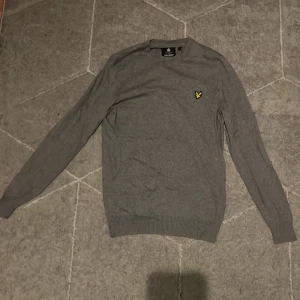 Grå stickad tröja från Lyle & Scott - Snygg grå stickad tröja från Lyle & Scott med klassisk gul broderad fågel på bröstet. Tröjan har rund hals, ribbade muddar och långärmad passform. Perfekt för dig som gillar stilrena och enkla plagg med en ikonisk detalj.