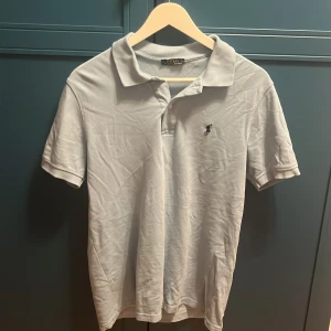Ljusblå pikétröja från Polo Ralph Lauren - Snygg vit pikétröja från Polo Ralph Lauren med klassisk krage och korta ärmar. Tröjan har den ikoniska broderade loggan på bröstet och är tillverkad i mjuk bomull. Perfekt för en clean och stilren look. Säljs pga att jag vuxit ur den. Original pris 1200