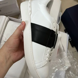 Valentino Open sneakers vit/svart - Snygga Valentino Open sneakers i vitt skinn med bred svart läderdetalj över mitten. Klassisk rund tå, platt sula och diskreta nitar baktill. Perfekta för dig som gillar stilrena och lyxiga sneakers med en twist.