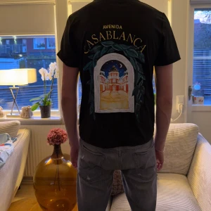 Svart Casablanca t-shirt med tryck - Svart t-shirt från Casablanca med färgstarkt grafiskt tryck på ryggen och mindre tryck på bröstet. Rund hals och korta ärmar. Trycket visar en nattlig scen med byggnader och gröna blad. Perfekt för dig som gillar coola prints och streetwear.