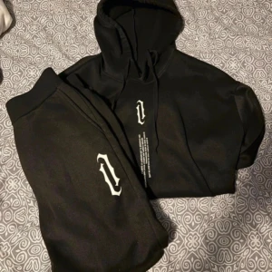 Svart hoodie och byxor med tryck från OCEANSAPART - Svart hoodie och byxor från OCEANSAPART med vit tryckt logga på bröstet och textdetalj under. Tröjan har huva med snörning och ribbade muddar vid ärmslut och nederkant. Perfekt för chill eller streetwear-stil.