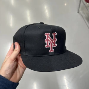 Svart NY keps med rosa brodyr - Snygg svart keps med rakt brätte och broderad NY-logga i rött framtill. Undersidan av skärmen är grön för extra stilpoäng. Klassisk snapback-modell som passar till streetwear och sportiga outfits.