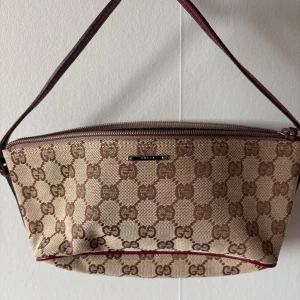 Beige och brun axelväska från Gucci - Snygg axelväska från Gucci i beige canvas med brunt GG-monogram och detaljer i vinrött skinn. Väskan har dragkedja upptill och ett klassiskt avlångt format. Perfekt för dig som gillar lyxiga accessoarer med ikonisk design.