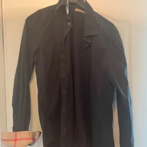 Svart skjorta från Burberry  - Snygg svart skjorta från Burberry i storlek L. Den har långa ärmar och klassiska rutiga detaljer på insidan av kragen och ärmsluten. Perfekt för både vardag och fest! Skjortan är bra skick och har knappar framtill. Hör av dig vid vid minsta fråga eller fundering! 😃
