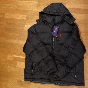 Svart dunjacka Polo Ralph Lauren XL - Svart dunjacka från Polo Ralph Lauren i storlek XL. Jackan har huva, två sidofickor med dragkedja och en bröstficka med dragkedja. Klassisk röd Polo-logga på bröstet och quiltad design. Perfekt för kalla dagar och riktigt snygg stil. Den sitter perfekt på mig och jag är 176