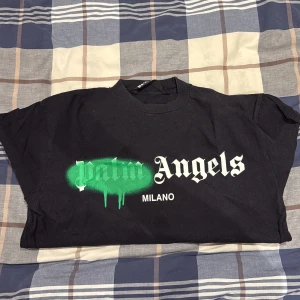 Palm Angels svart t-shirt Milano - Svart t-shirt från Palm Angels med Milano-tryck på bröstet. Framsidan har ett stort vitt och grönt grafiskt tryck där 'Palm' är spraymålat i grönt med dropp-effekt. Klassisk passform och rund hals. Perfekt för dig som gillar streetwear och statement-plagg.