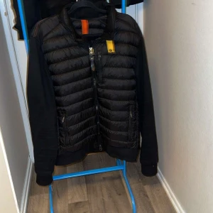 Svart pufferjacka från Parajumpers - Svart pufferjacka från Parajumpers med quiltad front och släta ärmar. Jackan har dragkedja, orange och gul detalj vid halsen samt patch på ärmen. Materialet är polyester och jackan har snygga fickor med dragkedja. Perfekt för dig som gillar streetwear och stilrena detaljer.