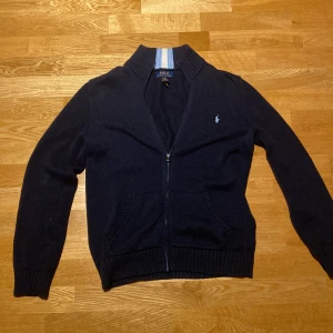 Mörkblå kofta från Polo Ralph Lauren - Snygg mörkblå kofta från Polo Ralph Lauren i stickad bomull. Tröjan har dragkedja framtill, ribbad krage med ljusblå och beige ränder, samt två fickor. Klassisk Polo-logga broderad på bröstet och långa ärmar. Perfekt för dig som gillar stilren design. Använd en gång så den är i jättebra skick. det är XL i barn så den passar till 175-180