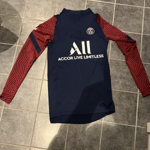 PSG långärmad träningströja Nike XS/S - Paris Saint-Germain långärmad träningströja från Nike i Dri-Fit material. Marinblå kropp med röda, randiga ärmar och PSG-logga på bröstet. Snygg halvdragkedja och vit tryckt sponsortext framtill. Perfekt för dig som vill ha en cool look på träningen. PASSAR BÄTTRE OM DU HAR STORLEK S