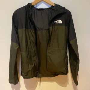 Svart och olivgrön vindjacka North Face - Snygg vindjacka från The North Face i svart och olivgrönt med huva och dragkedja framtill. Jackan har logga på bröstet och ryggen, samt praktiska fickor med dragkedja. Sitter som xs. Hör av dig för mer frågor💫 