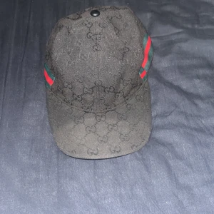 Svart Gucci keps med GG-mönster - Snygg svart keps från Gucci med klassiskt GG-mönster över hela kepsen och ikoniska gröna och röda ränder på sidorna. Kepsen har böjd skärm och justerbar rem baktill. Materialet är bomull och polyester, perfekt för dig som vill ha en lyxig streetstyle-look.