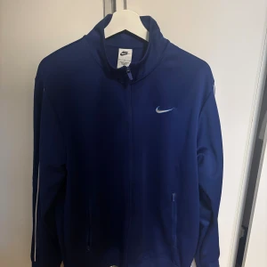 Blå vindjacka från Nike med dragkedja - Snygg blå vindjacka från Nike med vit logga på bröstet och ryggen. Jackan har hel dragkedja framtill, ståkrage och vita detaljer längs ärmarna. Perfekt för dig som gillar sportig stil och vill ha något bekvämt och stilrent.
