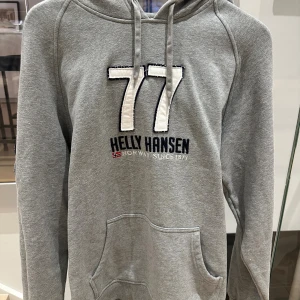 Grå hoodie Helly Hansen med tryck - Grå hoodie från Helly Hansen med stort vitt 77-tryck på bröstet och broderad logga. Tröjan har huva med snörning och en klassisk magficka. Mjuk bomullsmix som är skön att bära och passar perfekt för chill dagar. Snygg sportig stil med raglanärmar.