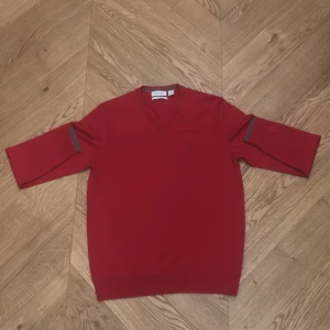 Röd merinotröja från Calvin Klein - Snygg röd tröja från Calvin Klein i extra fin merinoull. V-ringad med diskret ck-logo på bröstet och gråa ränder vid ärmsluten. Tröjan har trekvartsärm och en clean, modern look som passar perfekt till jeans eller chinos.