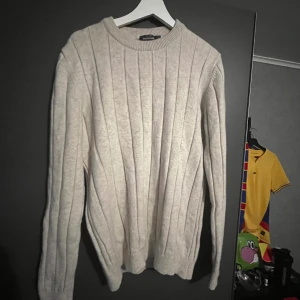 Ribbad beige stickad tröja Dressman - Säljer en beige stickad tröja från Dressman i storlek M. Tröjan har ett ribbat mönster med vertikala ränder och rund hals. Materialet är mjukt och passar perfekt när det är lite kyligare ute. Långa ärmar och klassisk passform.