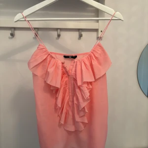 Rosa volang topp från H&M - Supersöt rosa volang topp från H&M med tunna axelband och volangdetaljer framtill.Toppen har en luftig känsla och är perfekt för varma dagar. Den är ärmlös och har en feminin v-ringning med flera lager volanger som ger extra liv till looken.