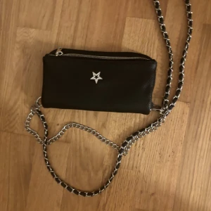 Svart axelväska med kedja från Gina Tricot - Svart axelväska i skinnimitation från Gina Tricot med silverfärgad kedja som axelrem. Väskan har en dekorativ stjärna med små stenar på framsidan och en praktisk dragkedja upptill. Perfekt storlek för mobilen och det viktigaste.