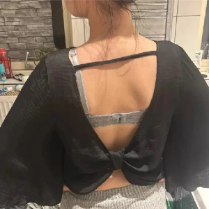 Festblus💕 - Säljer en svart blus med flowiga trekvartsärmar och snygg öppen rygg med knyt-detalj. Materialet är lätt och har en lite glansig finish som ger en cool vibe. Perfekt för dig som gillar statement-plagg med unik design. Framsidan är enkel medan ryggen har en twist.