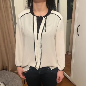 Vit blus med svarta detaljer från Zara - Snygg vit blus från Zara Basic med svarta detaljer längs krage, knytband och dekorativa sömmar. Långärmad och med lätt loose passform. Materialet känns som tunn och mjuk polyester, perfekt för en stilren look. Svarta prickar längs sömmarna ger extra edge. Passar även xs/s se bilderna🙂