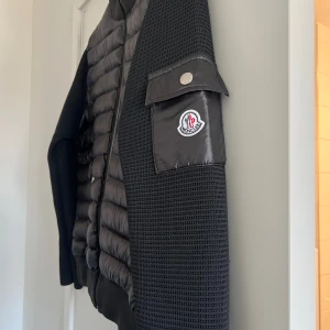 Svart Moncler pufferjacka med stickade ärmar - Svart pufferjacka från Moncler med stickade ärmar och quiltad front. Perfekt till höst/vinter. Jackan har två dragkedjefickor, hög krage och en snygg Moncler-logga på ärmen med fickdetalj. Materialmix av polyester och stickat ger en modern vibe. Väldigt liten i storlek passar min kille som är storlek M.  Jackan är endast provad ❗️osäker på Äkthet därav pris (finns inget kvitto)❗️