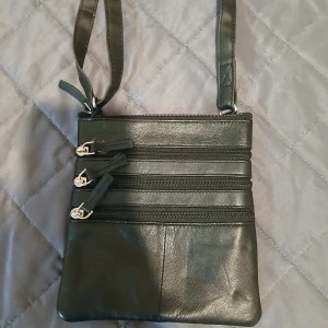 Svart axelväska i skinn med dragkedjor - Snygg svart axelväska i skinn med flera praktiska fack och dragkedjor framtill och baktill. Väskan har en lång, justerbar rem och stilren, slät design. Perfekt för dig som vill ha koll på dina grejer och gillar minimalistisk look.