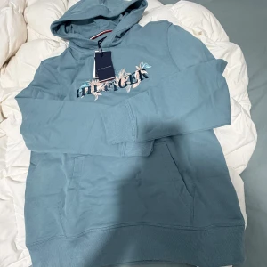 Blå hoodie från Tommy Hilfiger - Snygg ljusblå hoodie från Tommy Hilfiger med broderad logga och bladmönster på bröstet. Tröjan har huva, magficka och långa ärmar. Perfekt för dig som vill ha en chill och stilren look med en touch av färg.