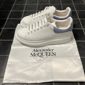 Alexander McQueen vita sneakers - Säljer ett par stilrena vita sneakers från Alexander McQueen med ljusblå mockadetalj på hälen och plattformssula i storlek 43. Skorna har rund tå, snörning och logotyp på både plös och häl. Perfekta för dig som gillar lyxiga och trendiga sneakers. Skorna är endast använda ett fåtal gånger, tyvärr ingår ingen skolåda. 