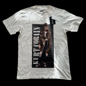 Vit Kurt Cobain t-shirt Carlings Couture - Vit t-shirt från Carlings Couture med stor tryckt bild på Kurt Cobain i leopardmönstrad jacka och texten 'KURT COBAIN' längs sidan. Klassisk rund hals och korta ärmar, tillverkad i mjuk bomull. Perfekt statement-plagg för dig som gillar musik och streetstyle.