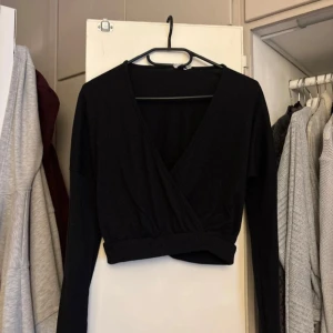 Svart omlott croppad topp - Svart långärmad croppad topp med omlott-design och djup v-ringning. Tunn ribbad struktur i tyget ger en snygg detalj. Perfekt att styla med höga jeans eller kjol för en trendig look.