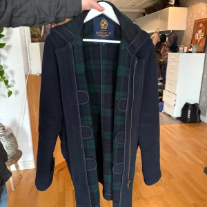 Mörkblå duffeljacka/rock från Bondelid - Säljer en mörkblå duffeljacka från Bondelid med huva och klassiska träknappar med snörning samt dragkedja. Insidan har ett grönt och svart rutigt foder som ger extra stilpoäng. Jackan har stora fickor framtill och snygga detaljer vid ärmsluten. Perfekt för dig som gillar preppy och tidlös look.