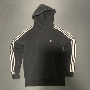 Svart Adidas hoodie med vita ränder - Svart hoodie från Adidas med klassiska vita tre ränder längs ärmarna och liten Adidas-logga på bröstet. Tröjan har huva, känguruficka och långa ärmar. Perfekt för en sportig och avslappnad stil.