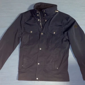 Mörkblå jacka från J.Lindeberg - Snygg mörkblå vindjacka från J.Lindeberg med hög krage, silverfärgade knappar och flera fickor framtill. Jackan är tillverkad i polyester och har en stilren look med diskret logopatch på ärmen. Perfekt för dig som gillar minimalistisk och sportig stil.