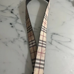 Rutig Burberry bälte i skinn - Snyggt Burberry bälte i klassiskt rutigt mönster med beige, svart och röda toner. Bältet är tillverkat i skinn och har en stilren form med metallspänne. Perfekt för att lyfta din outfit med en ikonisk detalj från Burberry.